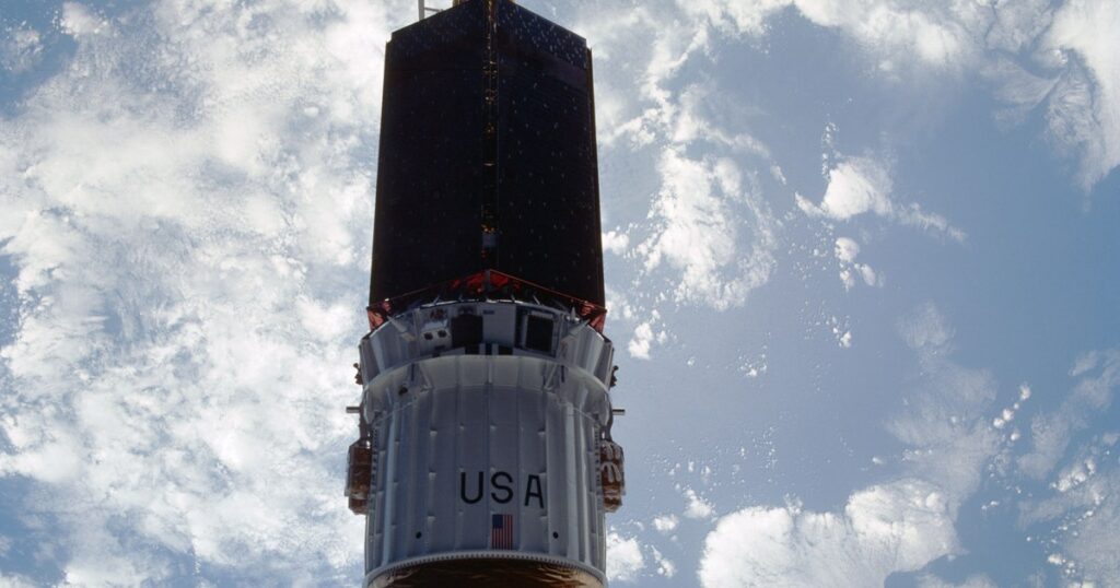 A tall cylindrical communications satellite with its upper-stage booster drifting away against a backdrop of white clouds over a blue ocean