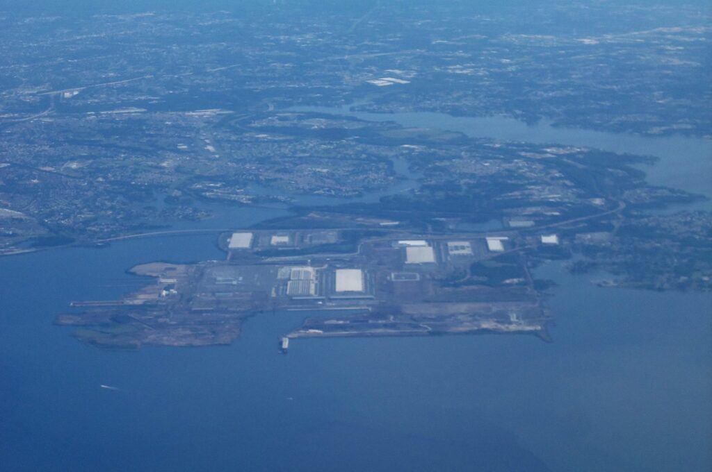 Aerial view of the Sparrows Point peninsula in Baltimore County, Maryland, looking north, photographed in June 2021: a flat, largely cleared waterfront site with large white warehouse roofs and docks, surrounded by Chesapeake Bay waters